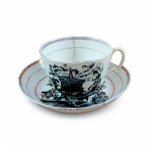 May include: A white teacup and saucer with a black and white transferware design depicting a scene with a carriage and figures. The saucer has a red rim.