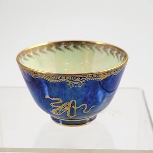 May include: A small, ornate blue bowl with gold accents. The bowl has a glossy finish with a gold rim and decorative gold patterns. The interior is a pale green color. A gold design is on the side of the bowl.