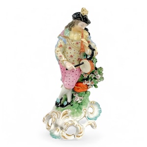 May include: A porcelain figurine of a musician playing a flute and a drum. The figure wears a black hat, a patterned jacket, and pink polka-dotted trousers. The base is decorated with flowers and gold accents.