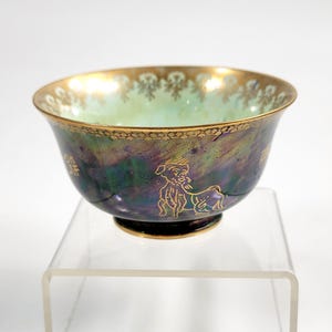 May include: An iridescent ceramic bowl with a gold rim and base. The bowl's exterior features a gold illustration of a mythical creature and gold Chinese characters. The interior has a light green hue.