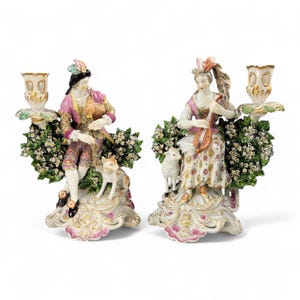 May include: Pair of porcelain candle holders featuring figures in 18th-century attire. One figure plays a bagpipe, the other a stringed instrument. Each is surrounded by floral arrangements and accompanied by a dog and a lamb. The candle holders have gold accents.