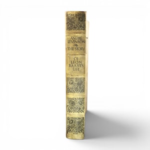 May include: A vintage book titled "Andre Levinson and The Story of Leon Bakst's Life." The spine is a light gold color with ornate, dark gold floral patterns. The book's title and author are printed in black.