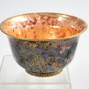 May include: A decorative bowl with a metallic finish. The interior has an orange and brown marbled effect, while the exterior is a dark blue with gold accents. Gold floral and geometric patterns adorn the bowl's rim and base.