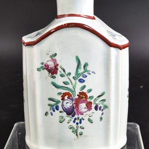 Puede incluir: Un bote de té de cerámica blanca con cuerpo de costillas verticales y cuello estrecho. La superficie presenta motivos florales pintados a mano en tonos rosa, morado, rojo, azul y verde, con un borde rojo oscuro a lo largo de los hombros.