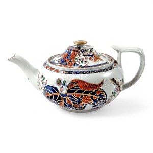 Puede incluir: Una tetera de porcelana blanca con un diseño floral azul, rojo y dorado. La tetera tiene un cuerpo redondo y un pico curvo.