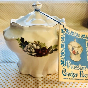 Op de afbeelding: Witte keramische koekjespot met een deksel en een bloemenmotief. De pot heeft een gouden rand en een blauw lint. Het etiket luidt "Prussian Cracker Bowl Approx. 2 1/2 lbs. Strawberry Preserves".