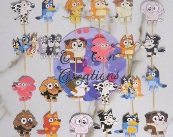May include: A set of 24 colorful cartoon dog cupcake toppers featuring characters from the show Bluey. The toppers are printed on cardstock and attached to wooden skewers. The characters include Bluey, Bingo, Bandit, Chilli, and more.