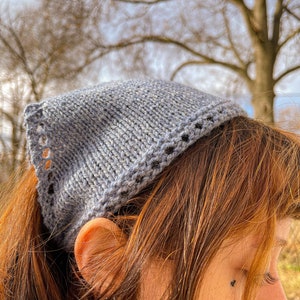 May include: A gray knitted headband with a decorative border. The headband is worn on a person's head with their hair pulled back in a ponytail.