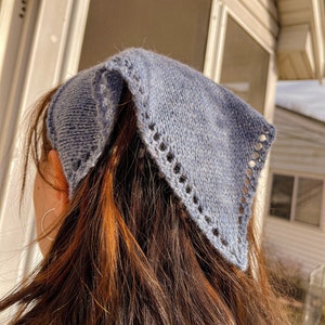 May include: A light blue knitted bandana with a delicate lace trim. The bandana is tied around the head of a person with long brown hair.