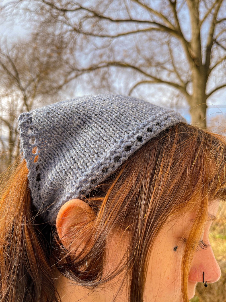 May include: A gray knitted headband with a decorative border. The headband is worn on a person's head with their hair pulled back in a ponytail.