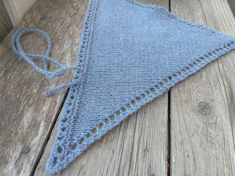May include: A blue knitted triangle shawl with a crocheted border. The shawl is lying on a wooden surface.