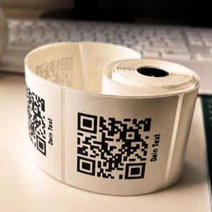 May include: A roll of white adhesive labels with black QR codes and the text "Dein Text" printed on them. The labels are rolled up, with some labels peeled back, revealing the QR code design.