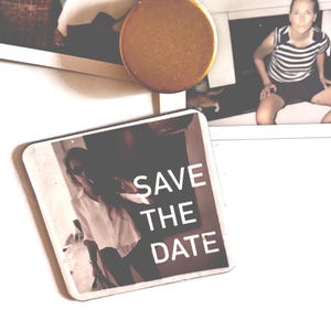 Könnte beinhalten: Ein quadratischer Magnet mit dem Text "SAVE THE DATE" in Weiß. Ein runder, brauner Magnet befindet sich darüber. Im Hintergrund ist ein Foto einer Person zu sehen.