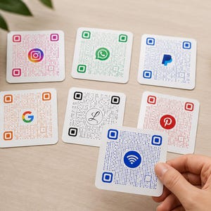 May include: A collection of white square QR code cards featuring social media and payment platform logos, including Instagram, WhatsApp, PayPal, Google, and Pinterest. One card is held by a hand, displaying a Wi-Fi symbol.