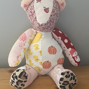Puede incluir: Un oso de peluche de patchwork confeccionado con varios patrones de tela, incluyendo fresas, patitos amarillos, calabazas naranjas, lunares rojos con un muñeco de nieve y estampado de leopardo. El oso tiene cara y orejas de color rosa claro.