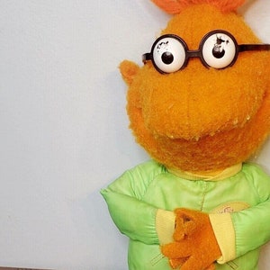 May include: Orange plush toy with red hair wearing a green and yellow jacket, blue jeans, and white trainers. The toy has large round eyes with black pupils and is wearing black framed glasses.