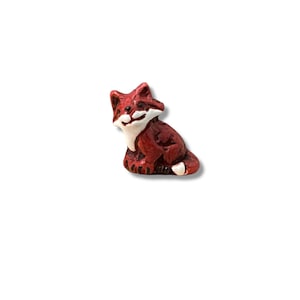 May include: A small, red fox figurine with white accents on its face and paws. The fox is in a sitting position, with its tail curled. The figurine is likely made of ceramic or a similar material.