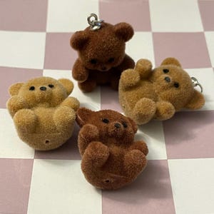 May include: Four small, fuzzy teddy bear charms in shades of brown and tan. Each bear has black bead eyes and a silver-colored metal loop for attaching to a keychain or other craft project. The bears are in various poses.