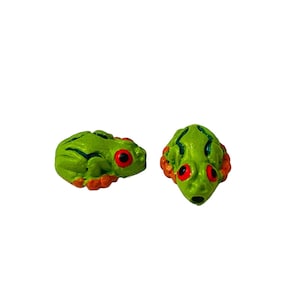 Puede incluir: Dos cuentas verdes con forma de rana con detalles en naranja y azul. Las ranas tienen ojos rojos y miran en direcciones diferentes.