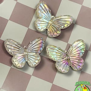 May include: Three iridescent butterfly-shaped beads with detailed wing patterns. The beads have a pearlescent sheen, reflecting a spectrum of colors. The background is a pink and white checkered pattern. The logo for "BANANARAMA BEADS" is in the bottom right corner.