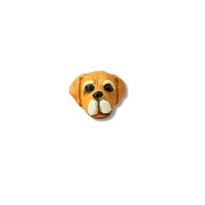 May include: A small, handcrafted dog head figurine. The dog has a light brown coat, black eyes, and a white muzzle. The figurine is set against a plain white background.