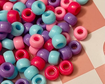 Puede incluir: Un montón de cuentas de plástico de colores en rosa, azul, morado y azul claro. Las cuentas son redondas y tienen un agujero en el centro. Están dispuestas sobre una superficie a cuadros rosa y blanca.