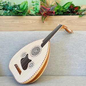 Puede incluir: Un oud de color claro, un instrumento de cuerda de Oriente Medio, descansa sobre una superficie gris. El cuerpo en forma de pera presenta intrincados agujeros de sonido y una tapa armónica marrón oscuro. El mástil y las clavijas son de madera. El instrumento está rodeado de vegetación.