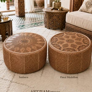 May include: Two round tan leather ottomans featuring intricate embossed patterns. The left ottoman displays a sunburst design, while the right ottoman features a floral medallion motif. Both pieces have detailed geometric side panels and text labels.