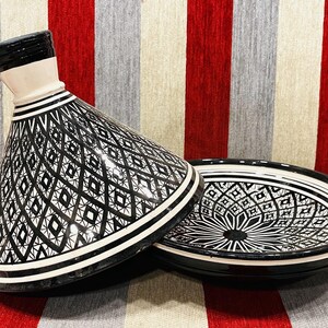 May include: A black and white ceramic tagine with a geometric pattern. The tagine has a lid and a base. The lid is slightly tilted to the side.
