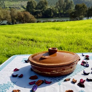 May include: A brown ceramic serving dish with a lid and a decorative design. The dish is sitting on a white tablecloth with a blue embroidered pattern and scattered flower petals.
