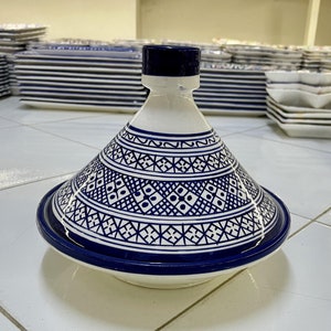 May include: A white ceramic tagine lid with a blue and white geometric pattern. The lid is round and has a pointed top. The lid is sitting on a white tiled surface.