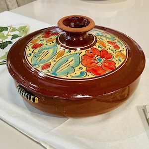 May include: A brown ceramic casserole dish with a lid. The lid has a floral design with red, yellow, blue, and green flowers. The dish has two black handles.