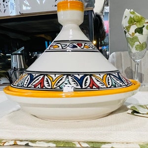 Op de afbeelding: Een witte keramische tajine met een gele rand en een kleurrijk bloemenpatroon. De tajine staat op een witte tafelkleed met een groen en wit patroon.