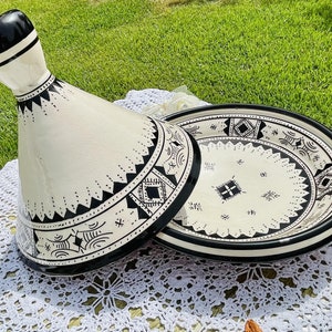 May include: A white ceramic tagine with a black rim and intricate black and white geometric designs. The tagine is open and the lid is resting on the side.
