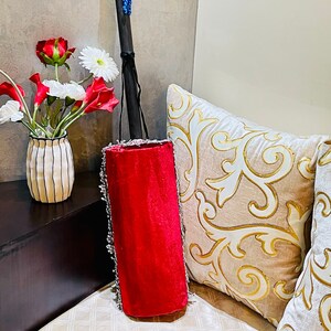 May include: A red velvet cylindrical cushion with silver trim and a black wooden handle with a blue tassel. The cushion is sitting on a white and gold patterned couch.