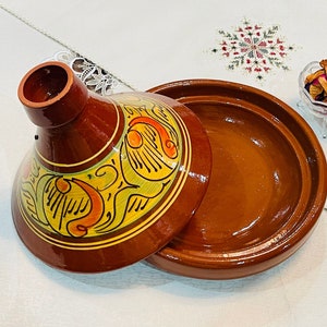 May include: A traditional Moroccan tagine cooking pot with a lid. The lid is decorated with colorful floral patterns in yellow, orange, and green. The pot is made of brown clay.