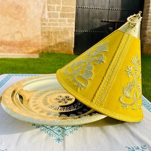 May include: A gold metal serving tray with an ornate design and a yellow fabric cover with embroidered details. The cover is shaped like a cone and has a handle on top.