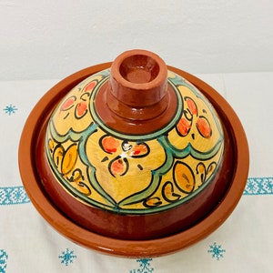 May include: A terracotta tagine with a lid, featuring a colorful floral design in shades of yellow, orange, green, and blue.