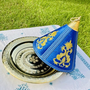 May include: A gold metal serving tray with intricate designs sits on a white tablecloth with blue embroidery. A blue velvet cone-shaped cover with gold embroidery sits on top of the tray.