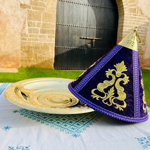 May include: A gold serving tray with intricate designs sits on a white tablecloth with a blue pattern. A purple velvet cone-shaped lid with gold embroidery and a gold handle sits on the tray.