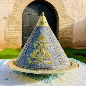 May include: A silver and gold Moroccan food cover with a blue and gold embroidered design. The cover is sitting on a white tablecloth with a blue pattern.
