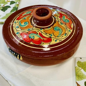 May include: A round, brown ceramic dish with a lid. The lid has a decorative design of yellow, orange, red, and blue floral patterns. The dish is perfect for serving food.