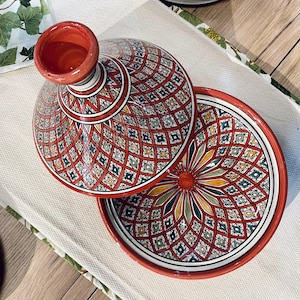 Puede incluir: Una tagine de cerámica roja y blanca con intrincados patrones geométricos. La tagine tiene una base redonda y una tapa abovedada con una pequeña abertura en la parte superior.