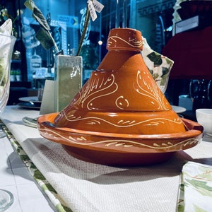 May include: A traditional Moroccan tagine cooking pot with a brown clay body and white decorative designs. The tagine is on a white tablecloth with a green and white patterned border.