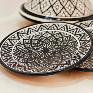 May include: A set of two white ceramic plates with black geometric designs. The plates have a raised rim and a center design that resembles a flower.