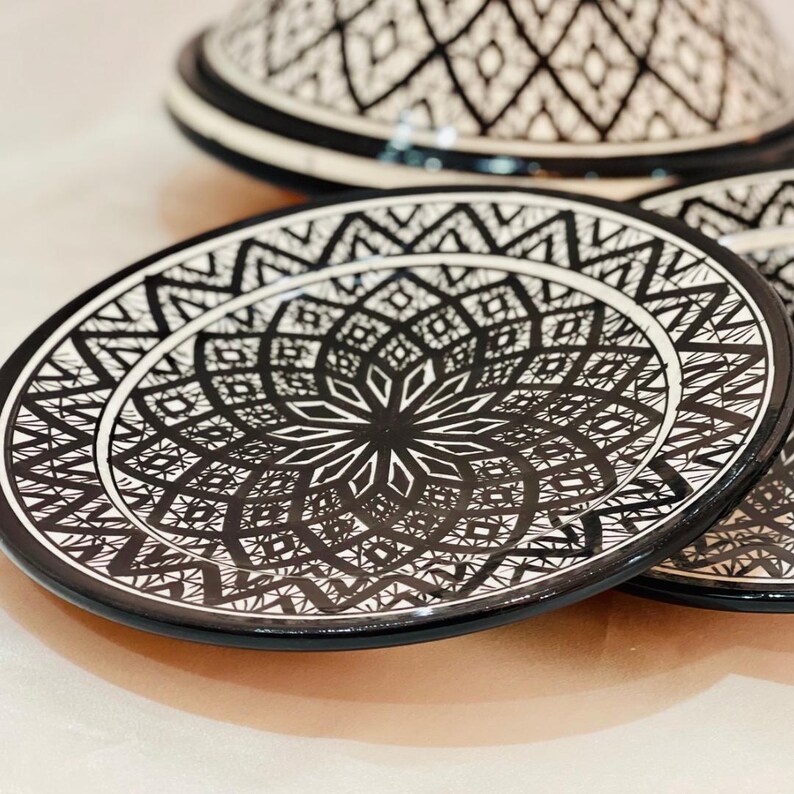 May include: A set of two white ceramic plates with black geometric designs. The plates have a raised rim and a center design that resembles a flower.