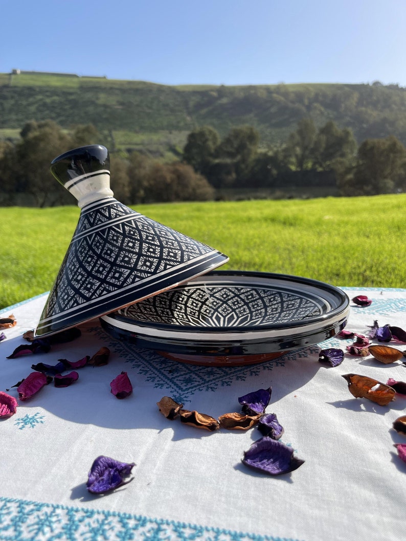Może przedstawiać: Czarno-biała ceramiczna tagine z geometrycznym wzorem. Tagine jest otwarta, a pokrywka leży z boku. Tagine stoi na białym obrusie z rozrzuconymi płatkami kwiat&oacute;w.
