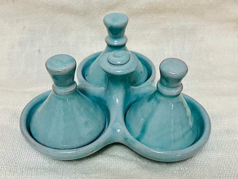 May include: A set of three teal ceramic condiment dishes with lids. The dishes are arranged in a triangular shape on a base.