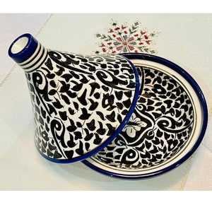 Könnte beinhalten: Eine weiße Keramik-Tagine mit einem schwarzen und blauen floralen Design. Die Tagine hat einen runden Boden und einen konischen Deckel.