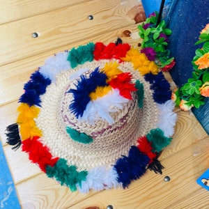 May include: A straw hat with a wide brim and colorful pom-poms around the edge. The pom-poms are in shades of red, yellow, blue, green, and white.